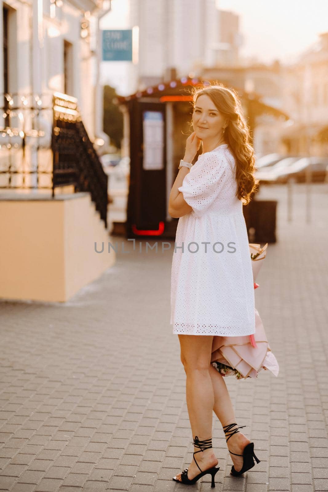 A happy woman in a white dress at sunset with a bouquet of flowers in the city.