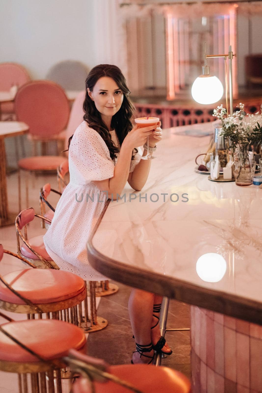 A girl in a white dress is sitting at the bar in a cafe and drinking a cocktail.