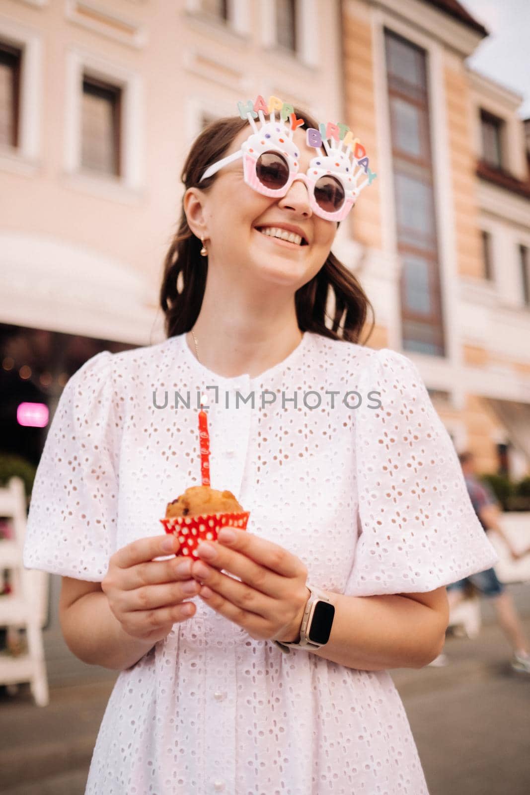 A beautiful happy woman in a white dress holds a cake in her hands on the street of the city celebrating her birthday.