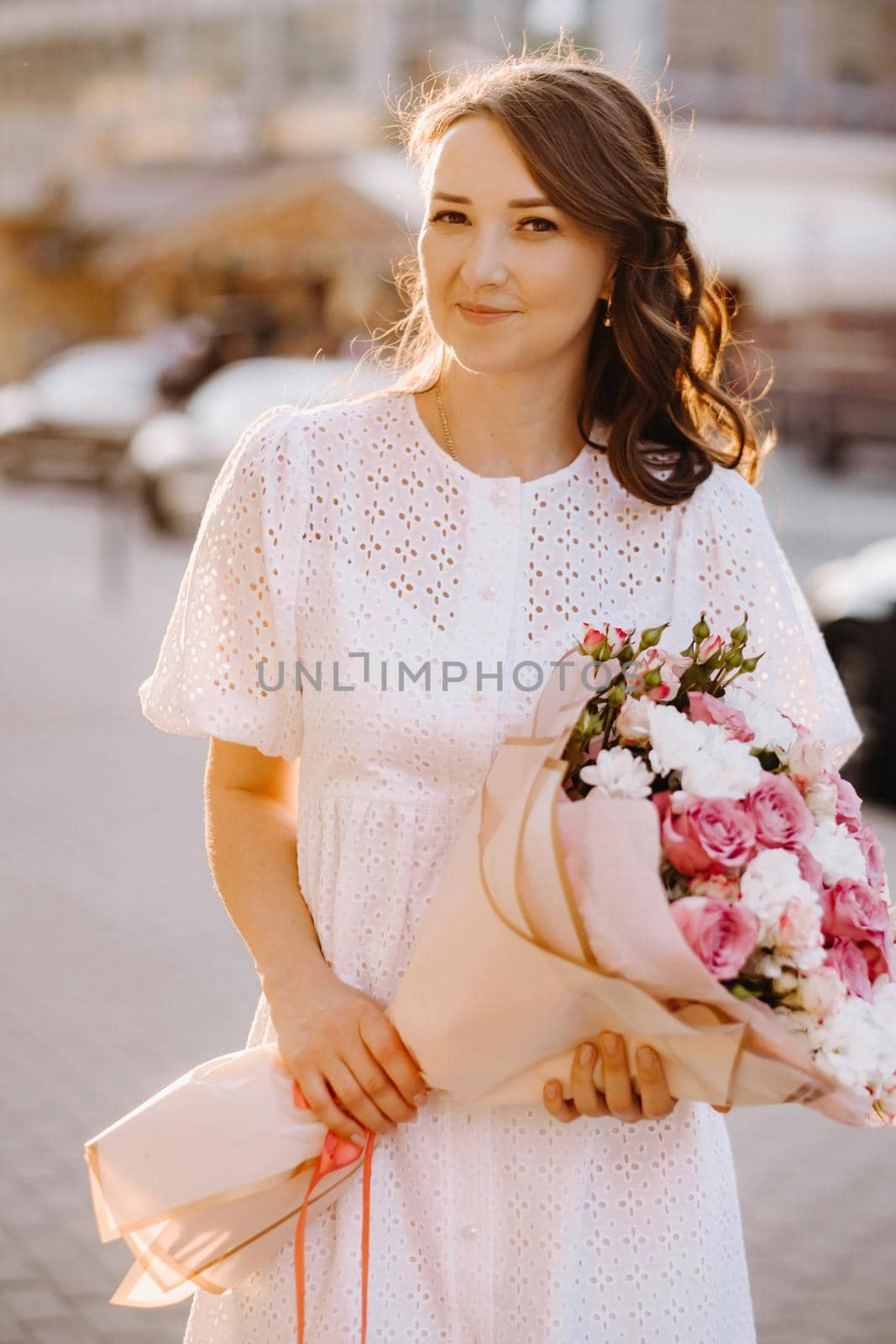 A happy woman in a white dress at sunset with a bouquet of flowers in the city.