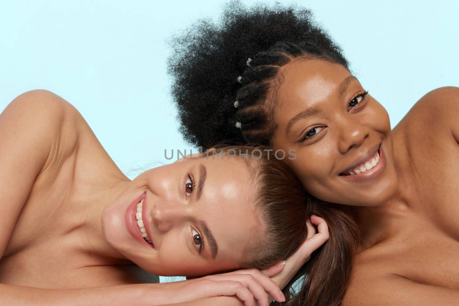 Young beautiful caucasian and african women posing lying on top of each other, face to face. Lifestyle of different nationalities, people concept. Looking at camera.