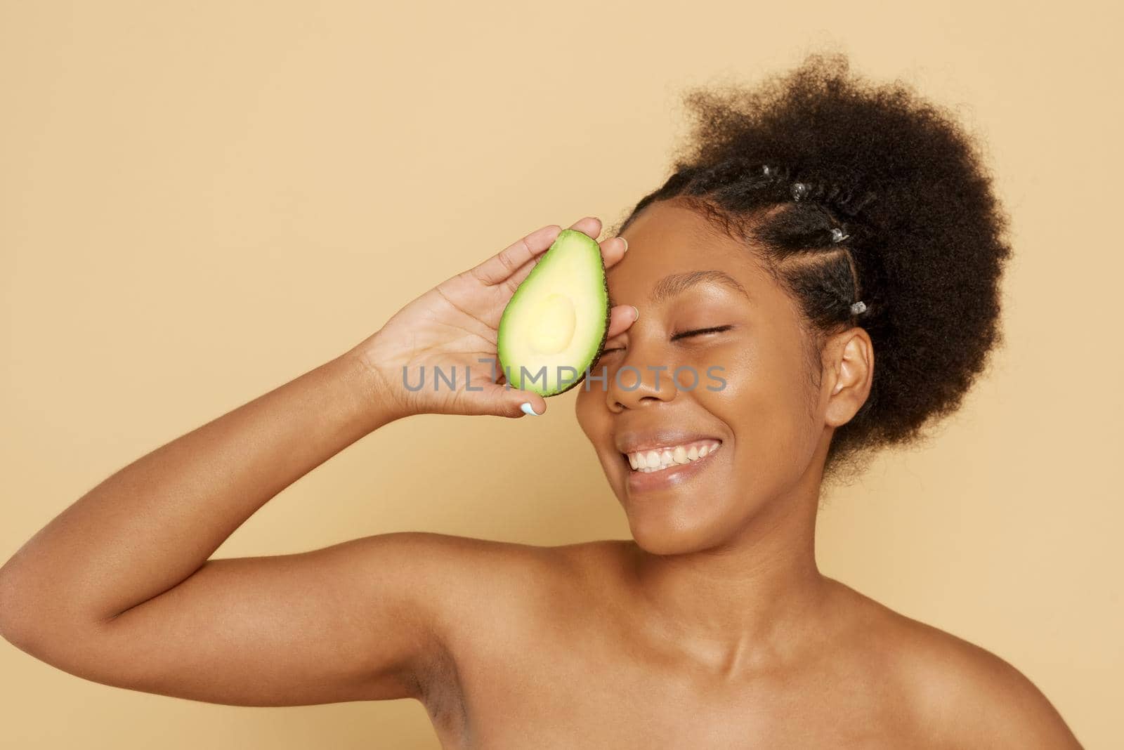 Portrait of a beautiful young African woman holding an avocado and smiling against a beige background. The concept of proper nutrition, skin care, spa and cosmetology.