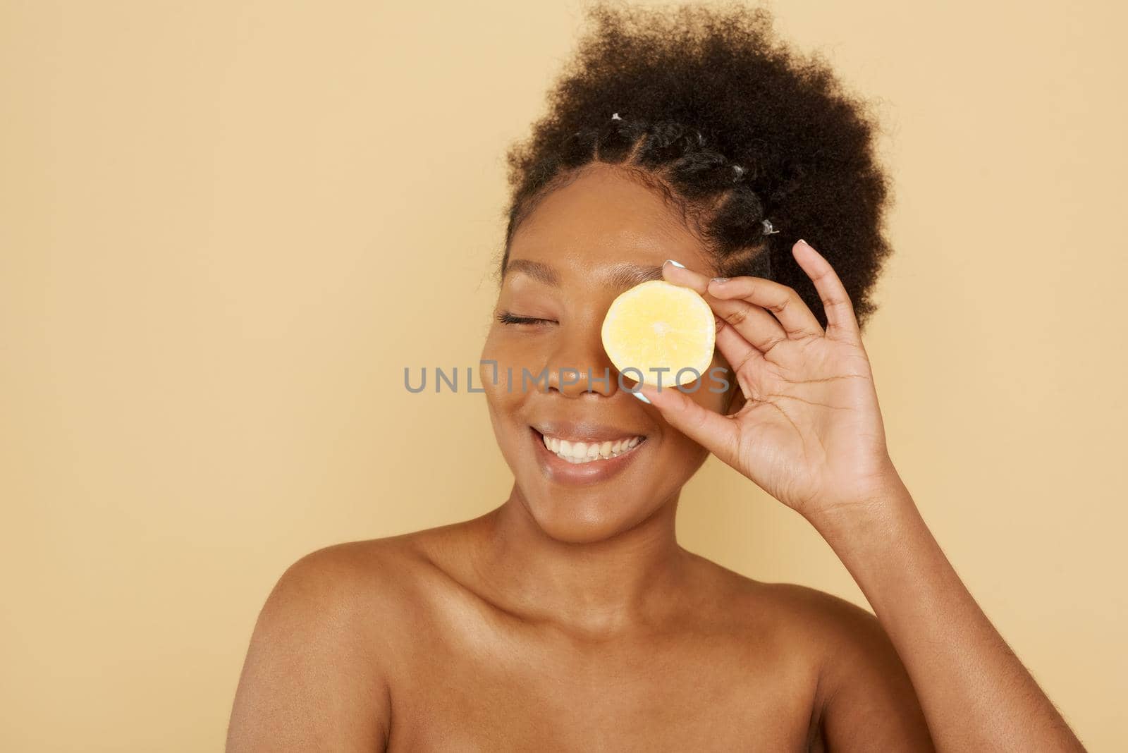 Beautiful afro american female model holding half a lemon in front of her face on a beige background. The concept of caring cosmetics with vitamin c, skin care and spa