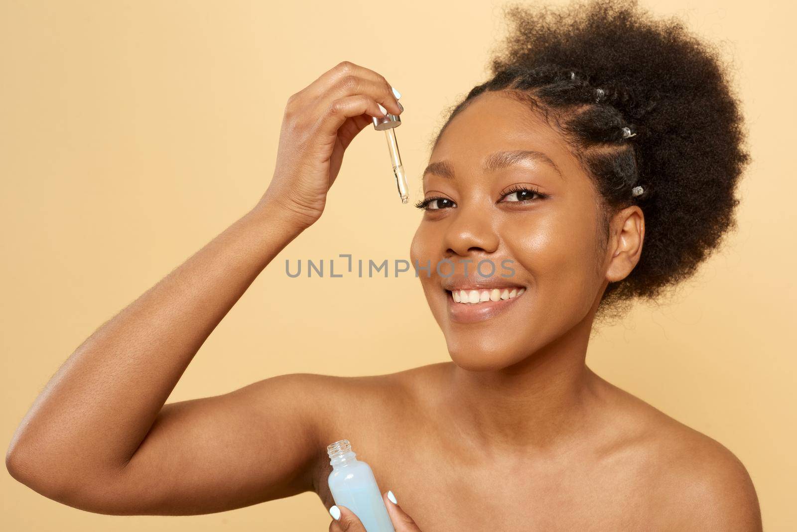 The concept of beauty, cosmetics and skin care. Portrait of happy smiling young african american woman with bare shoulders applying facial serum or oil with pipette on face on beige background and looking at camera.