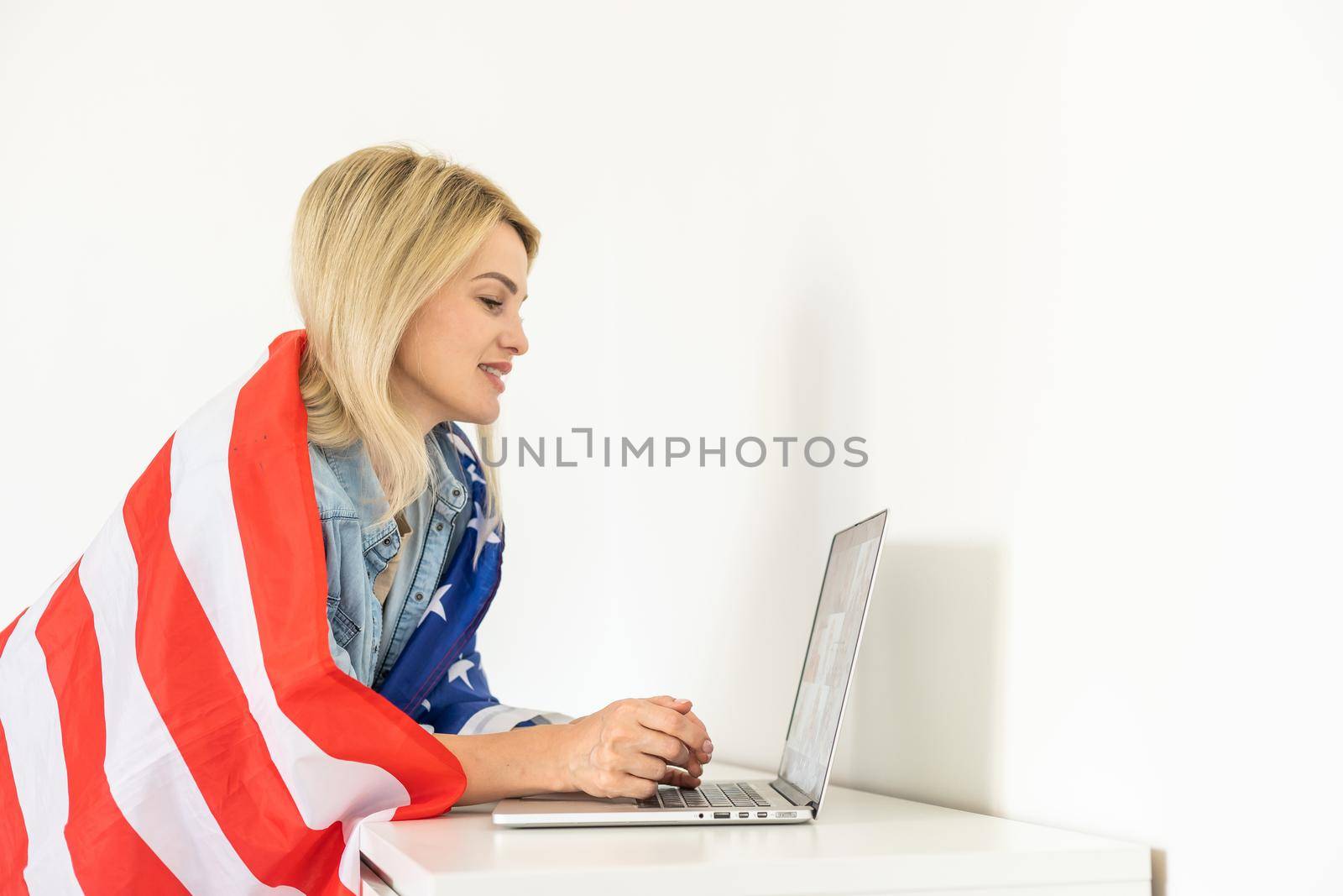 Happy woman employee sitting wrapped in USA flag, shouting for joy in office workplace, celebrating labor day or US Independence day. Indoor studio studio shot isolated on yellow background.