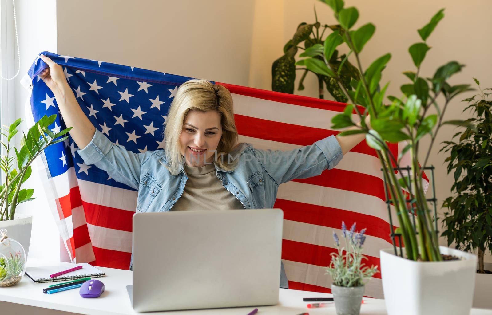 Happy young female student hold flag of USA, studying with laptop with blank screen in living room interior. International education at home, lesson remote, website due covid-19 quarantine.