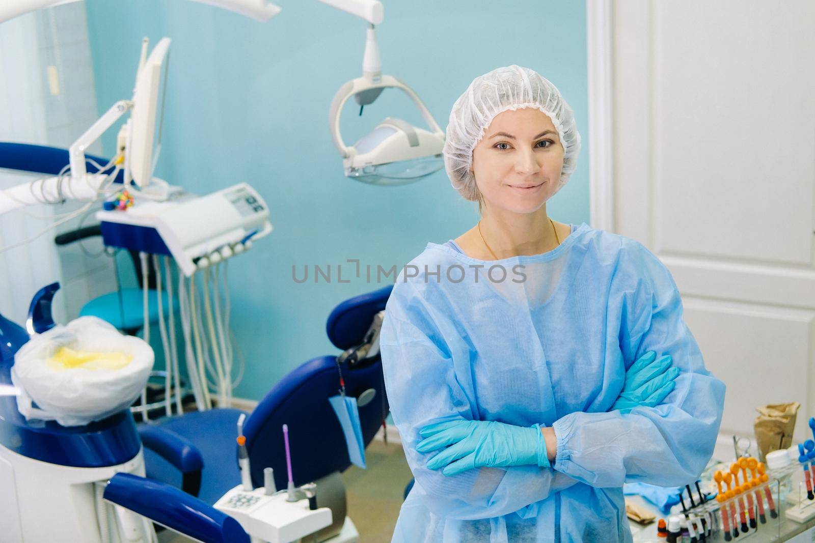 a female dentist wearing a medical mask and rubber gloves poses for the camera and folds her arms in her office by Lobachad