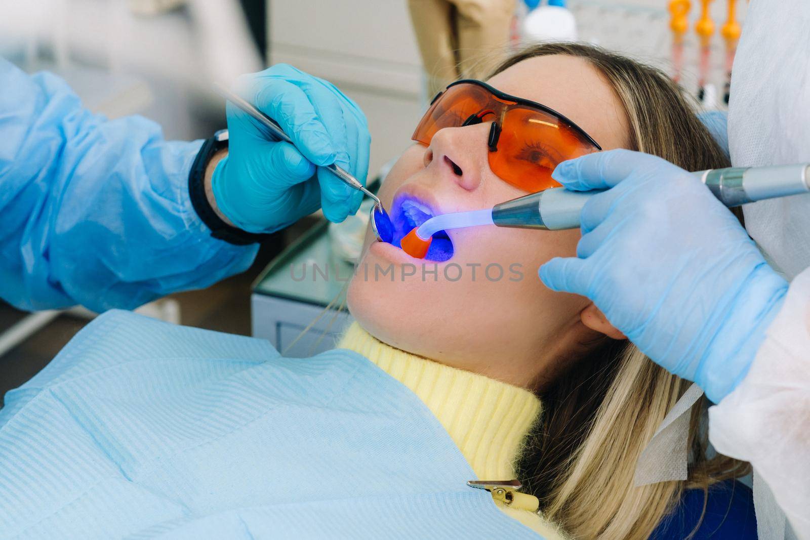 A young beautiful girl in dental glasses treats her teeth at the dentist with ultraviolet light. filling of teeth by Lobachad