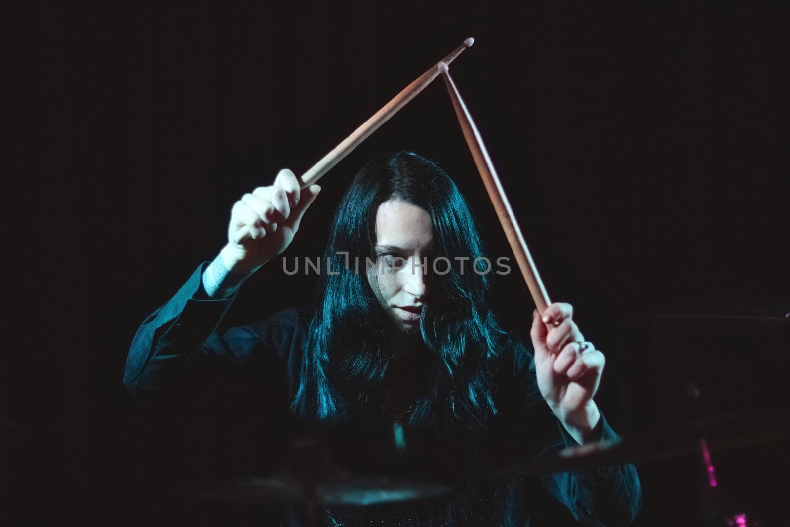 A female metal drummer with long hair holding up drumsticks behind the drumkit against a dark background by tennesseewitney