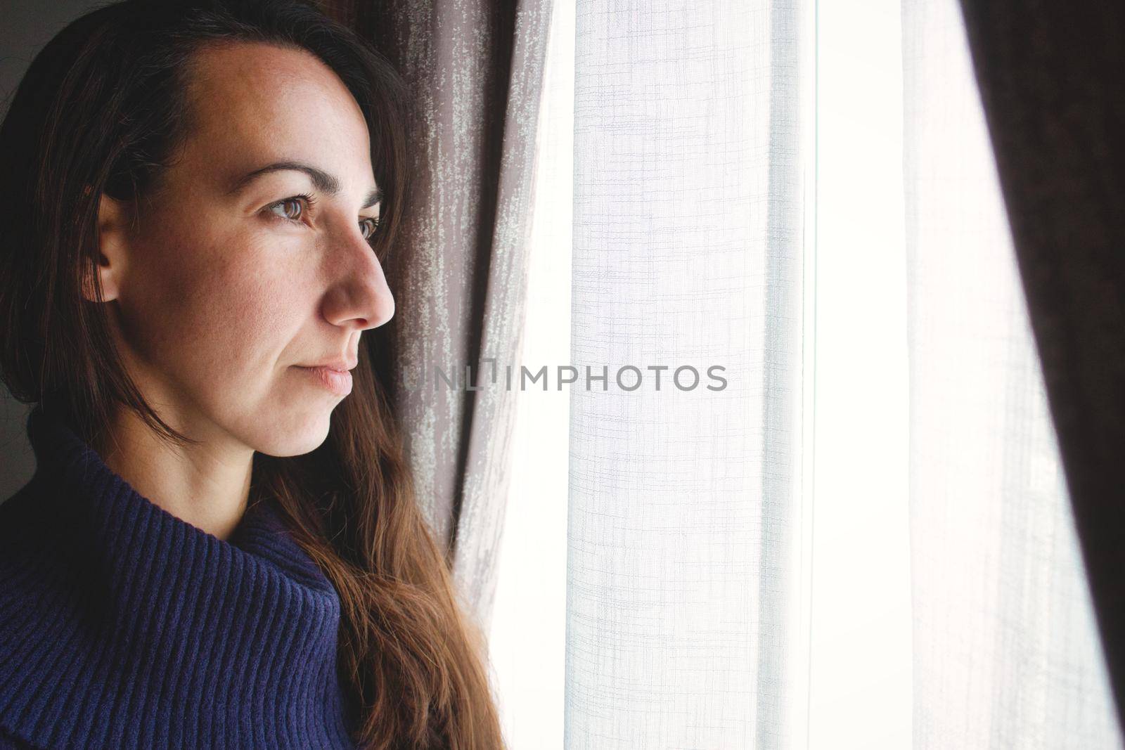 Young attractive woman looking pensively out of a window with curtain and glowing white light