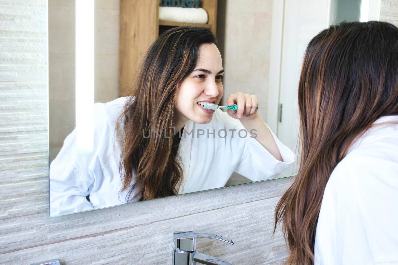 Lifestyle shot of attractive white Caucasian woman brushing her teeth in front of the bathroom mirror wearing a white robe