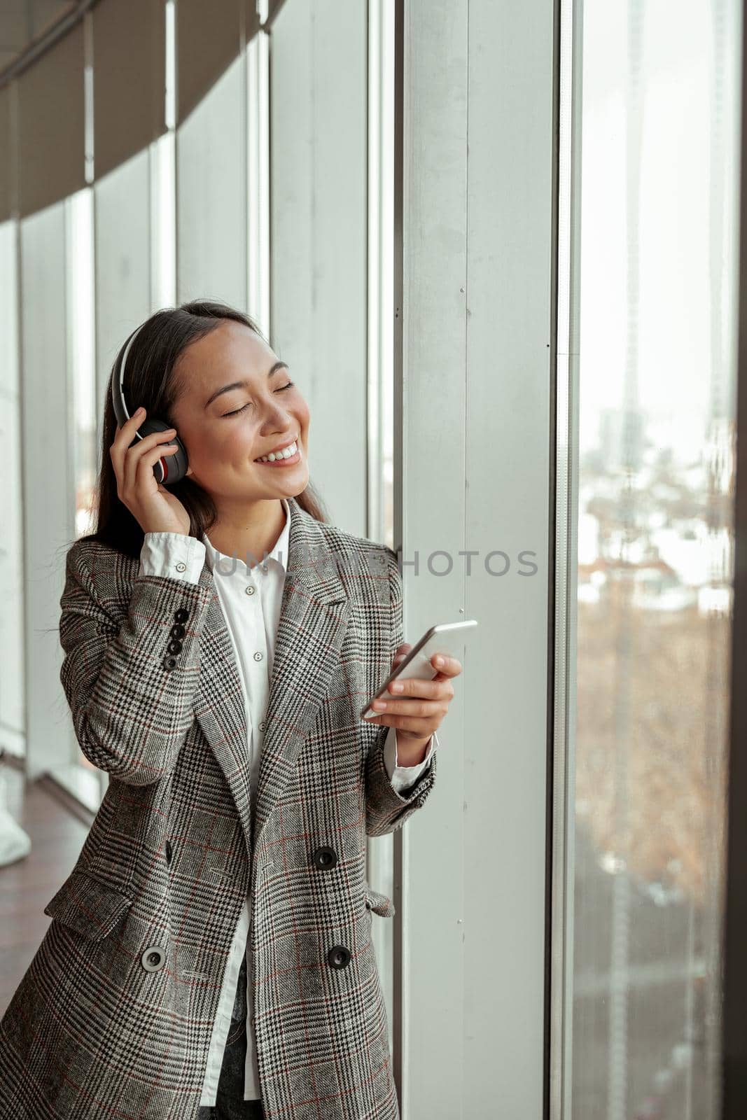Asian Business woman listening music in headphones standing near window in office during break by Yaroslav_astakhov