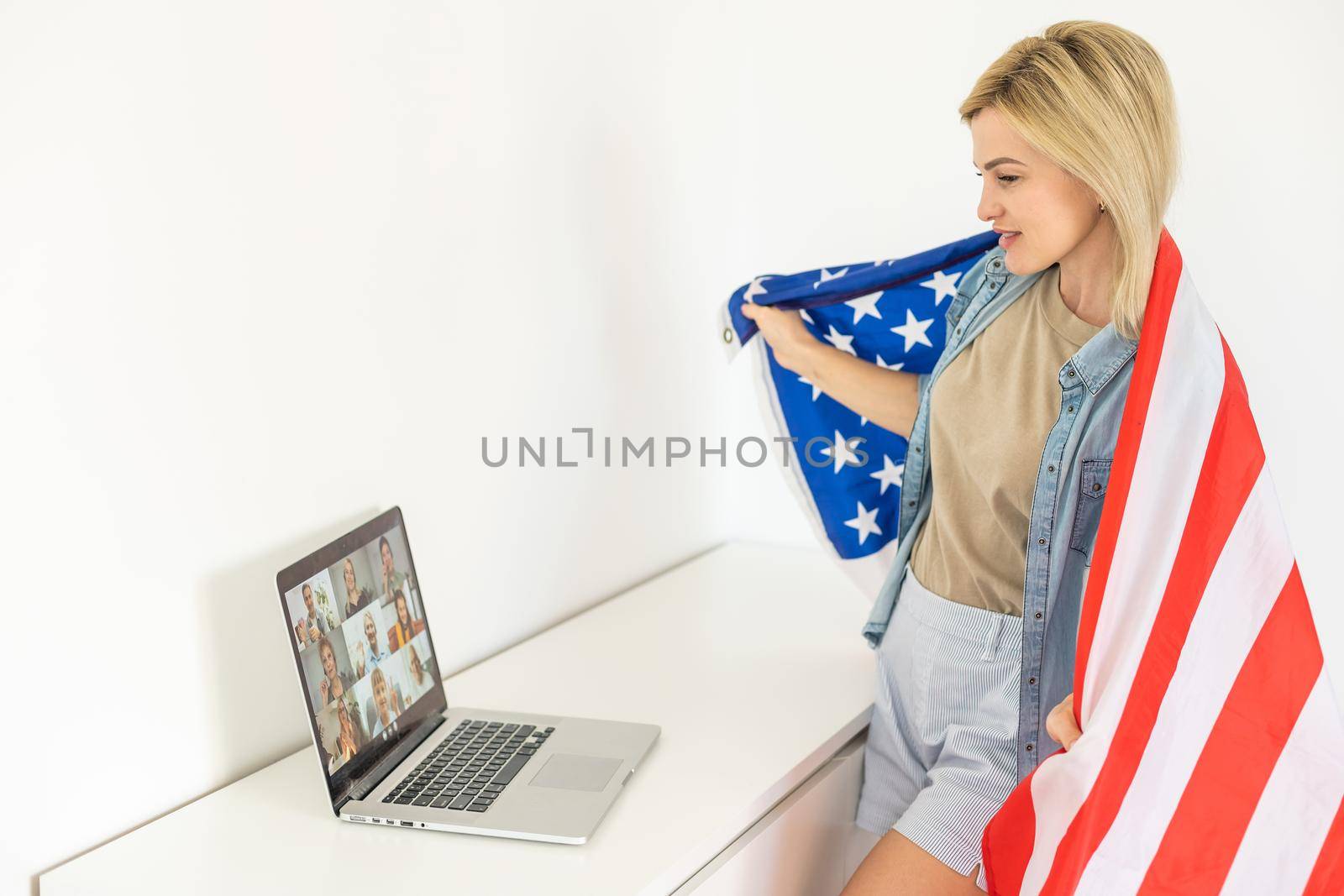 Happy woman employee sitting wrapped in USA flag, shouting for joy in office workplace, celebrating labor day or US Independence day. Indoor studio studio shot isolated on yellow background.