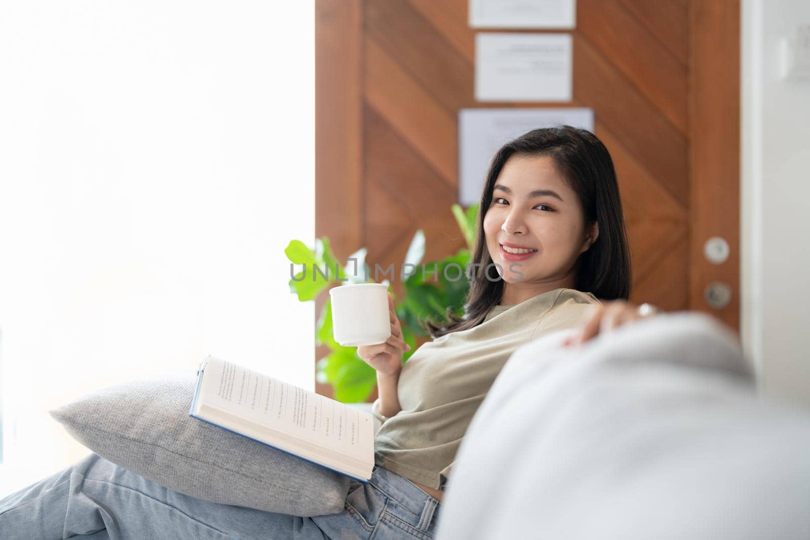 Smiling asian woman on couch reading a book looking at camera. coffee morning