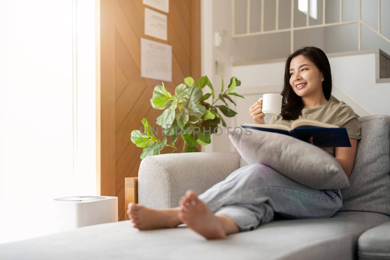 happy asian young woman sitting on sofa and reading book at home -people and leisure concept by nateemee