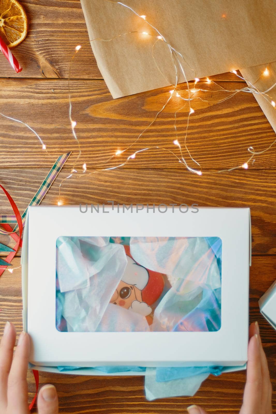 Box of Christmas gingerbread on wooden table. Women's hands reach for carton of cookies. Garland, wrapping paper and ribbons. New Year composition.