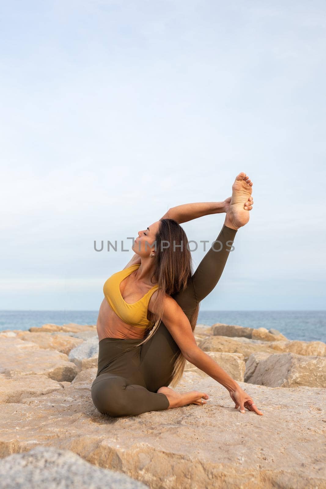 Flexible young woman doing Parivritta Kraunchasana yoga pose on a rock near the sea. Copy space. Vertical image. Healthy and active lifestyle concept.