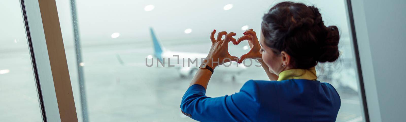 Female flight attendant making heart sign with hands while standing by the window and enjoying airfield view