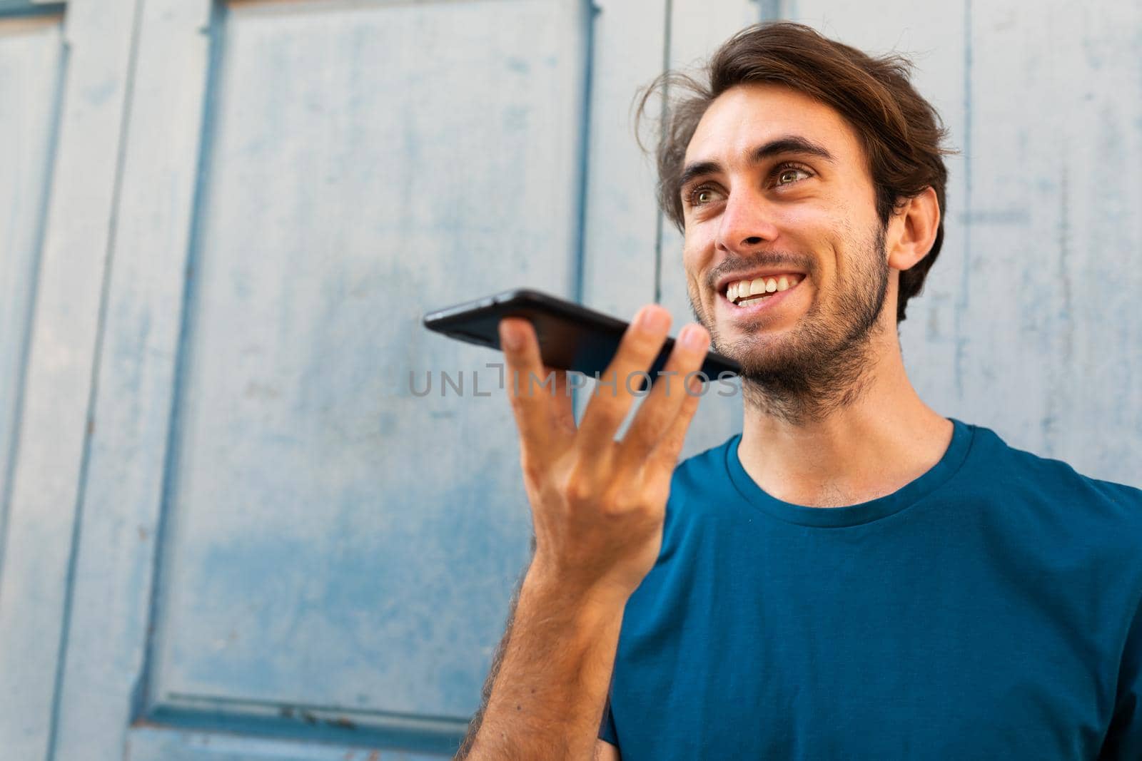 Young caucasian man sending voice message with mobile phone outdoors. Copy space. by Hoverstock