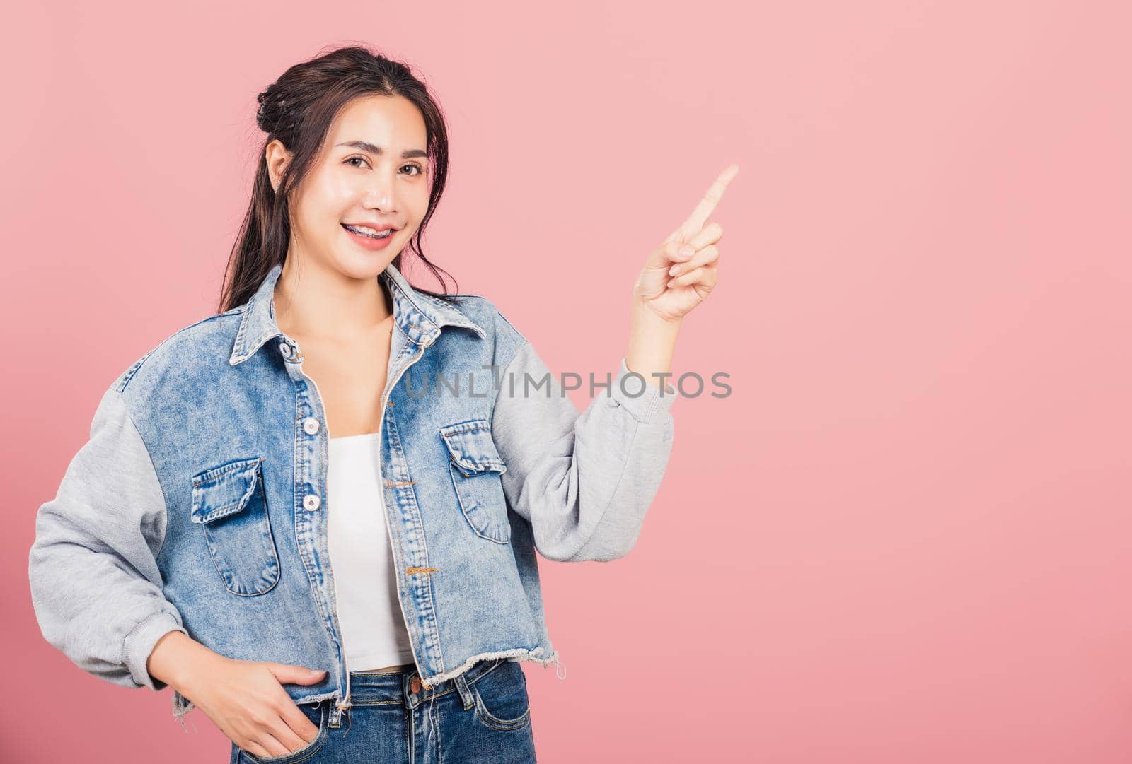 Happy Asian portrait beautiful cute young woman standing smiling indicate finger empty space looking to camera, studio shot isolated on pink background, Thai female pointing index out with copy space