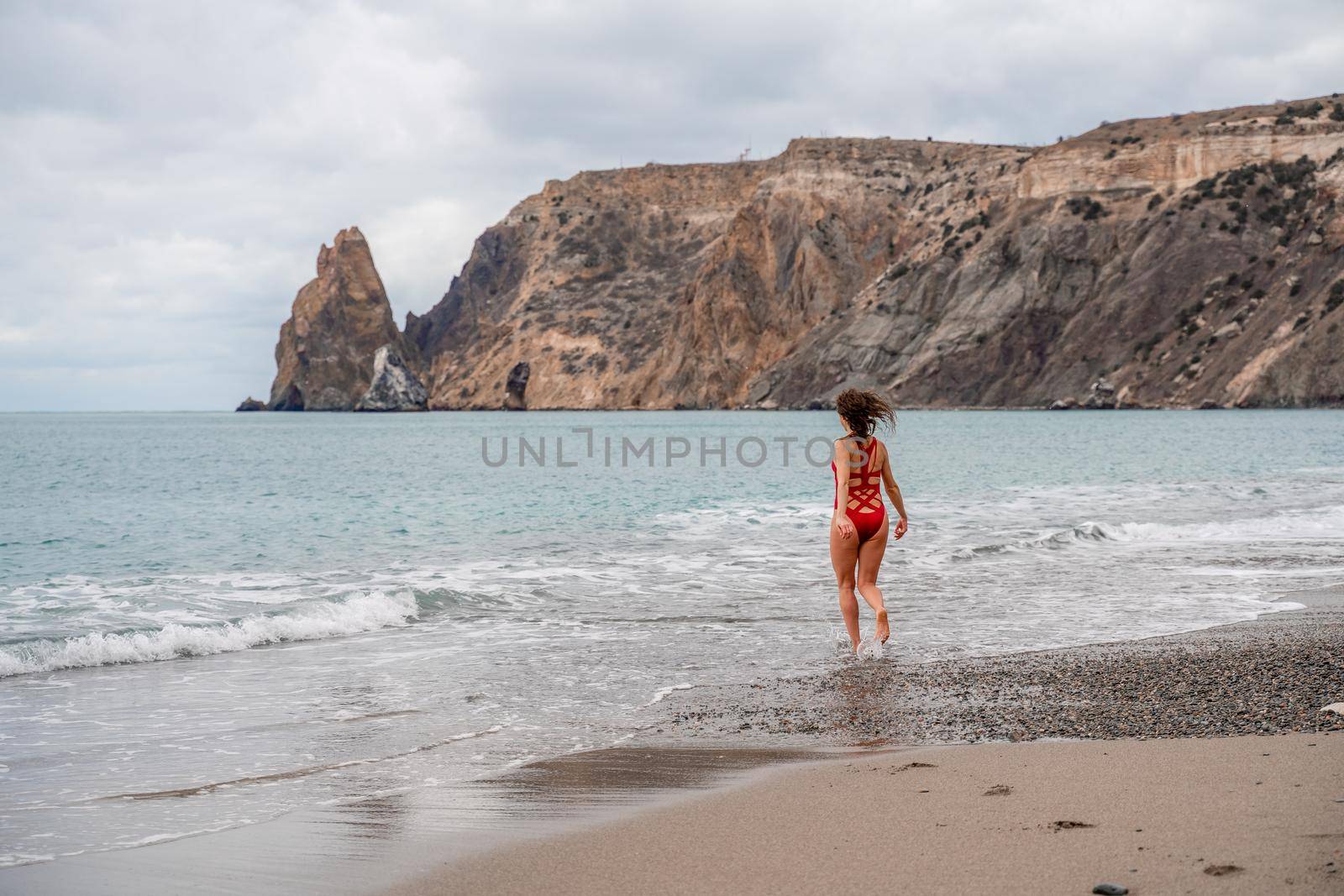 A beautiful and sexy brunette in a red swimsuit on a pebble beach, Running along the shore in the foam of the waves by Matiunina