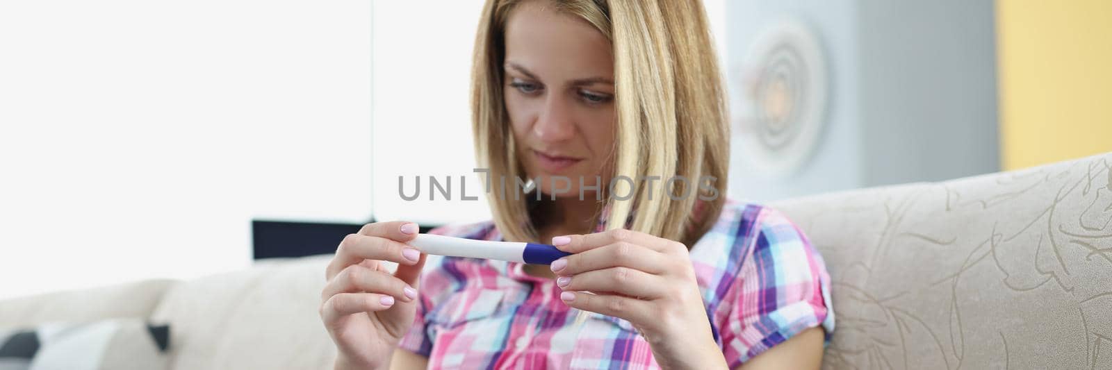 Portrait of young woman hold pregnancy test, happy female got big news. Blonde woman sit on sofa at home. Pregnancy, new period, life change, baby concept