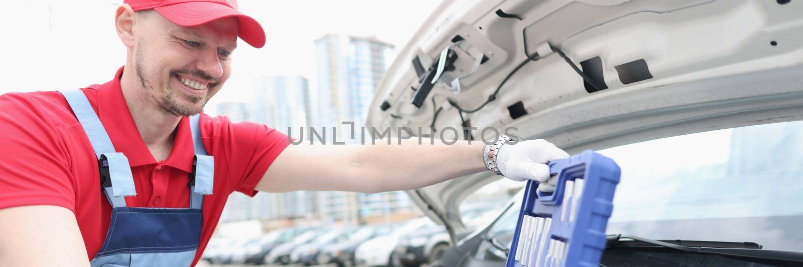 Portrait of automotive technician smiling and open kit full of equipments. Open car hood and need to fix problem of automobile. Car maintenance concept