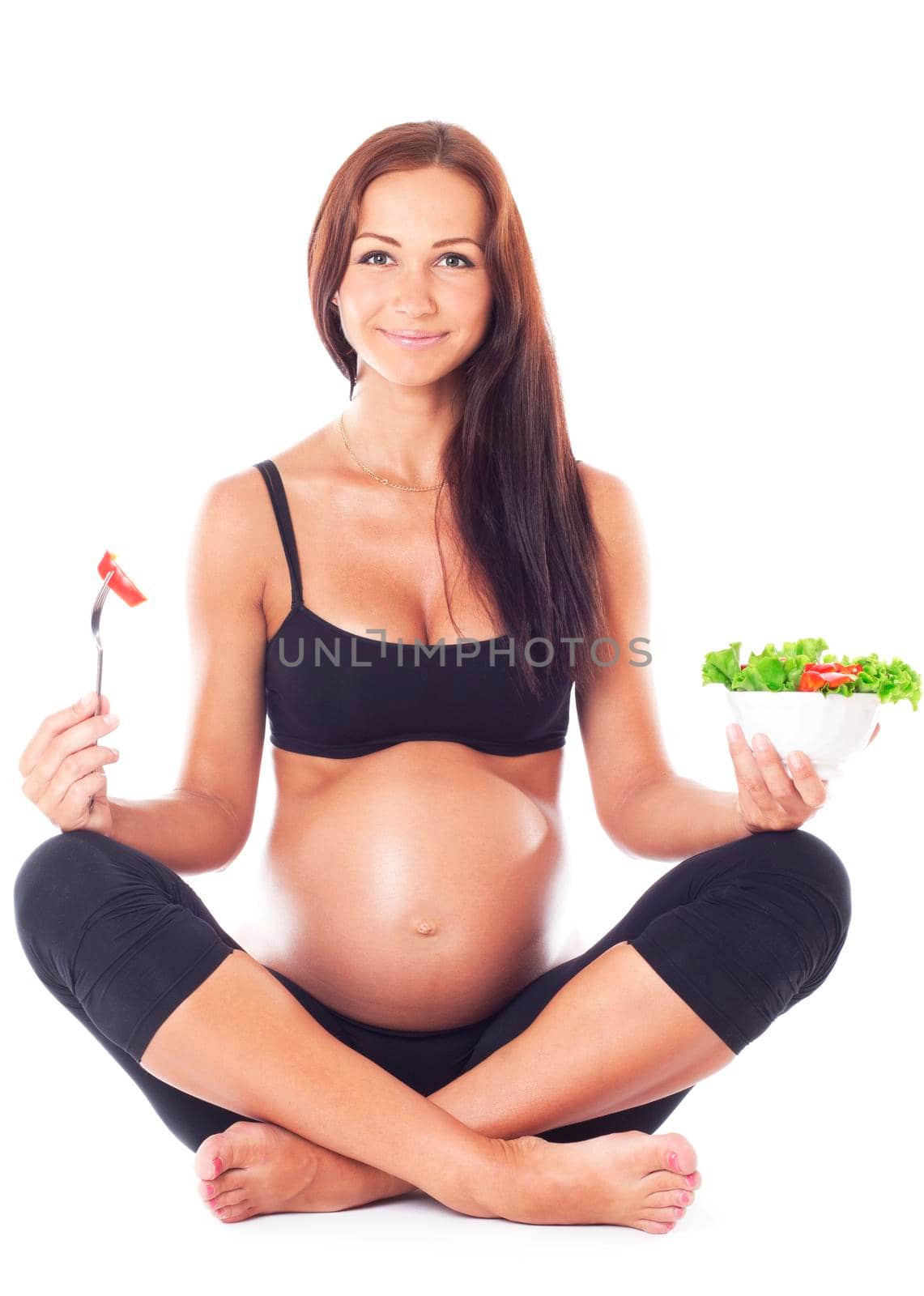 Pregnant woman sitting cross-legged and eating chopped salad, isolated on white.