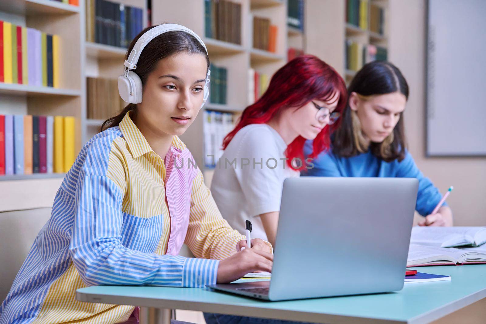 Group of teenage students study in school library classroom. In focus is teenager girl in headphones using laptop. High school, learning, education, knowledge, adolescence, people concept