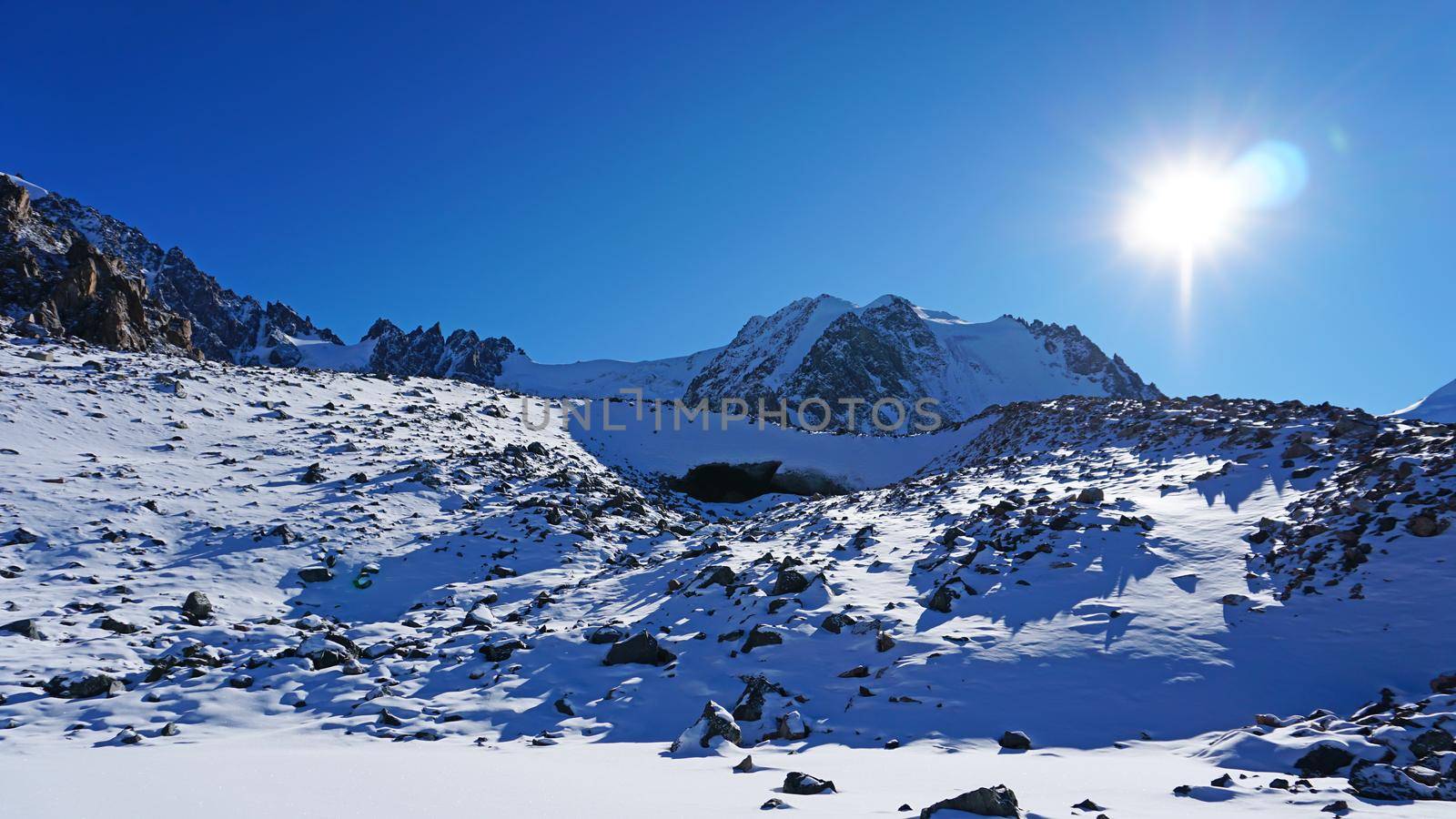 Entrance to an ice cave in the mountains. Tuyuk Su Glacier, all covered in white snow. You can see an ice grotto covered with ice and rocks. High rocks and mountains. The entrance looks like a dolphin