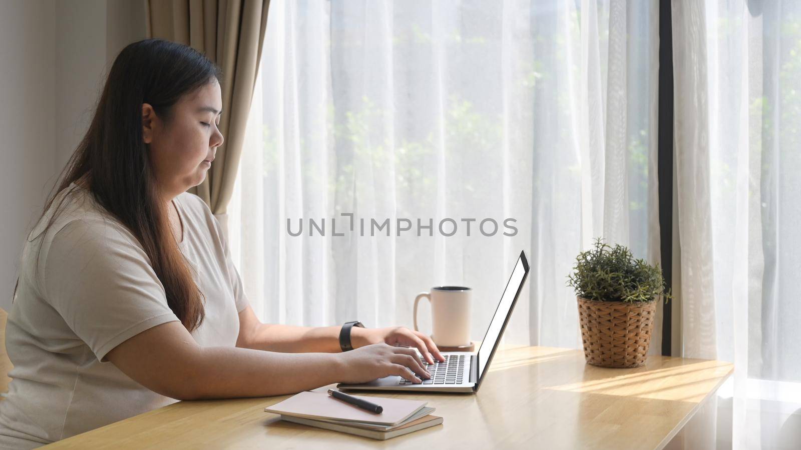 Young overweight woman freelancer using laptop while working remotely at home office.