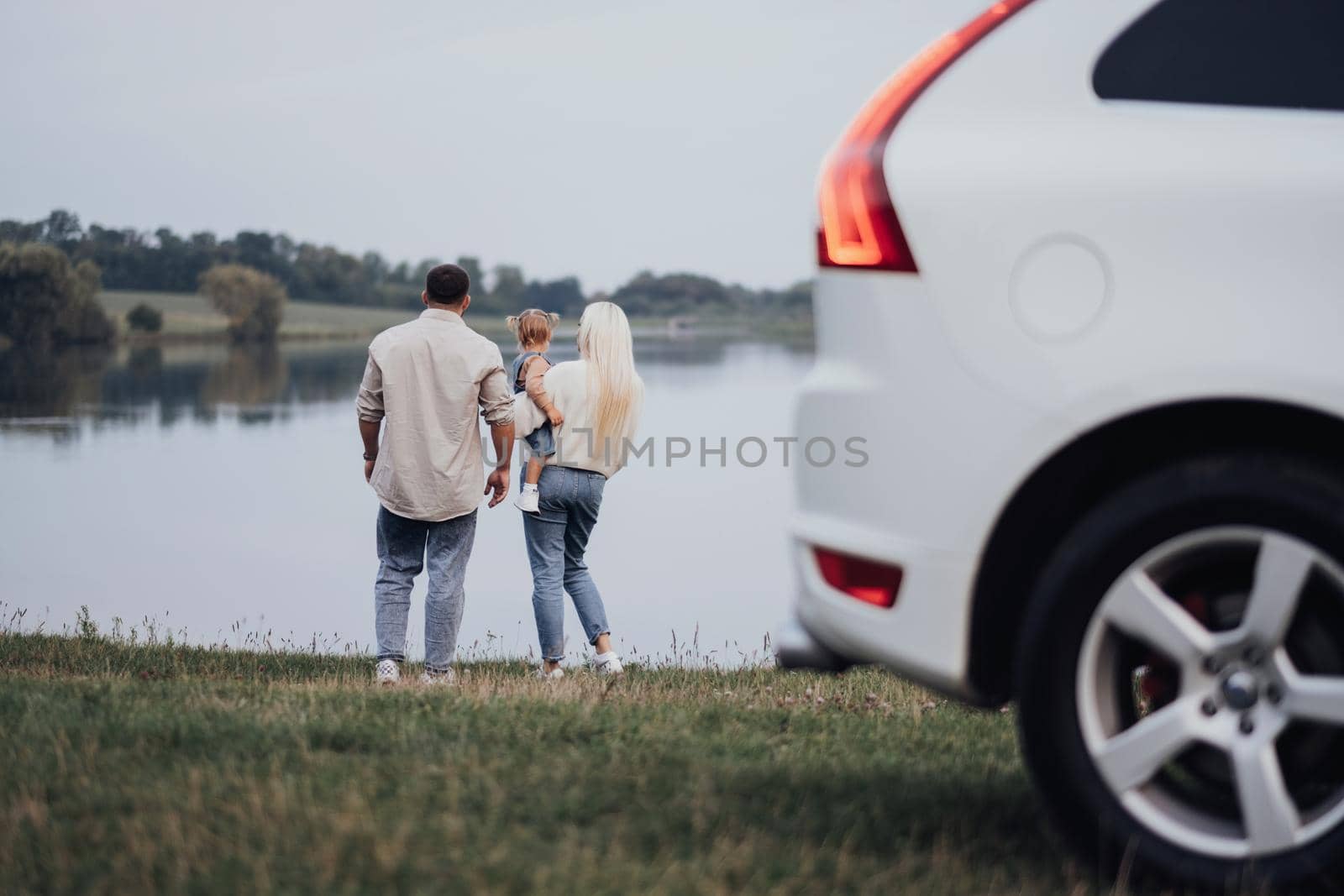 Young Mother and Father with Their Toddler Daughter Standing Outdoors Near the Lake, Concept of Family Road Trip on SUV Car