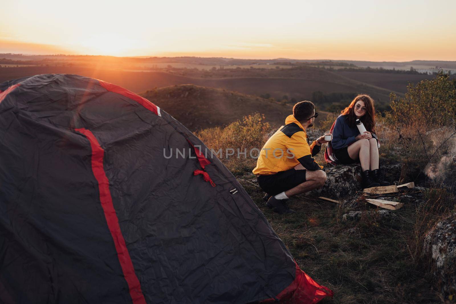 Two Young Travelers Man and Woman Sitting Near Campfire and Tent During Sunset Over Top of Hill