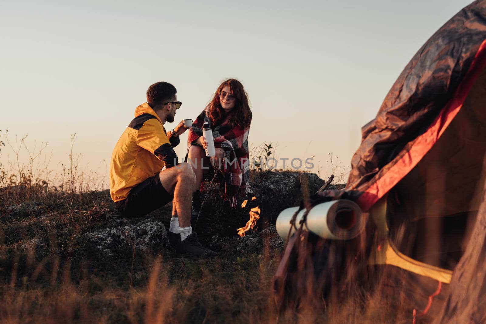 Two Young Travelers Man and Woman Sitting Near Campfire and Tent Outdoors During Sunset and Drinking Hot Tea from Thermos