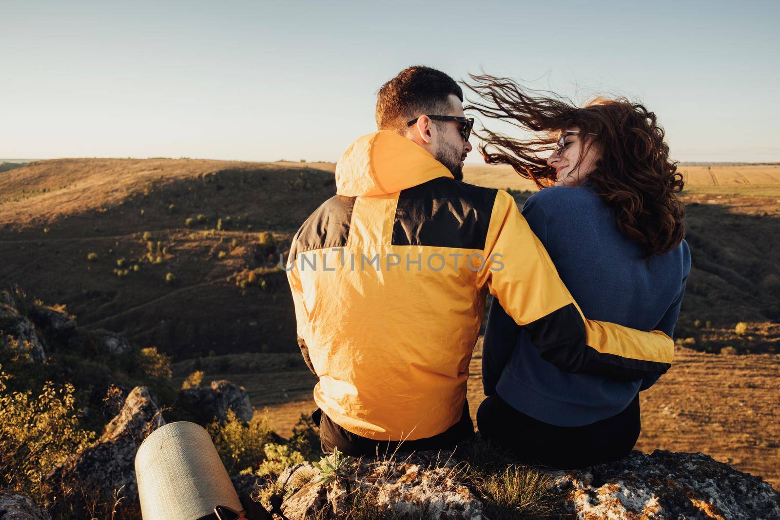 Back View of Traveler Couple Hugging and Sitting on Edge of Rock During Sunset, Wind Blowing Woman's Hair