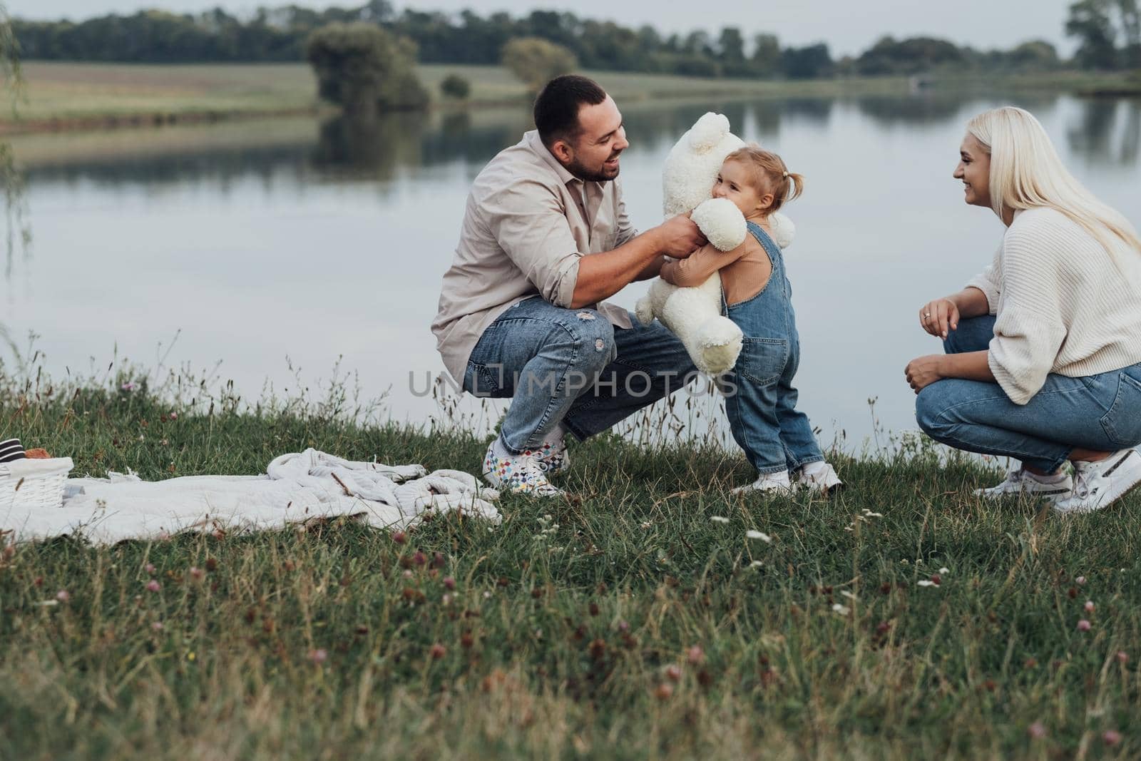 Father Gave His Child Soft Toy, Little Daughter Hugging Teddy Bear, Young Family Enjoying Picnic Outdoors Near Lake