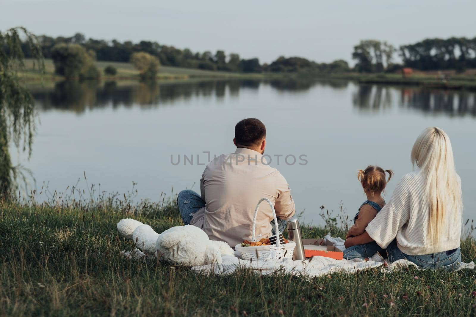 Back View of Young Family Having Picnic Time, Mom and Dad with Their Little Daughter Sitting Near the Lake Outside the City by Romvy