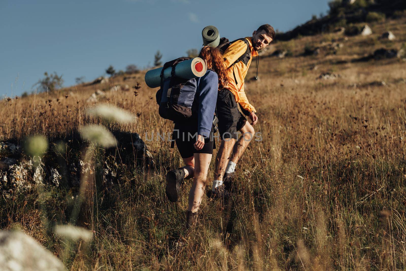 Two Travelers with Backpacks and Camping Mat on a Route, Man Helping Woman to Move Forward on Top of Hill During Sunset