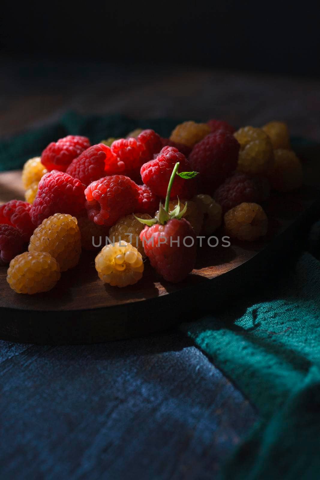 Red and yellow raspberry on wooden board on blue and green background, close up, selective focus low key.