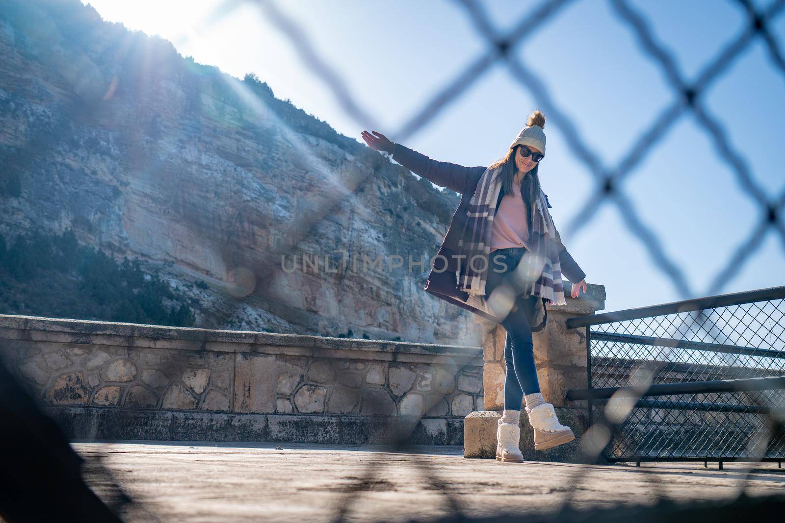 Young woman standing with outstretched hands wearing coat and hat behind a fence outdoors