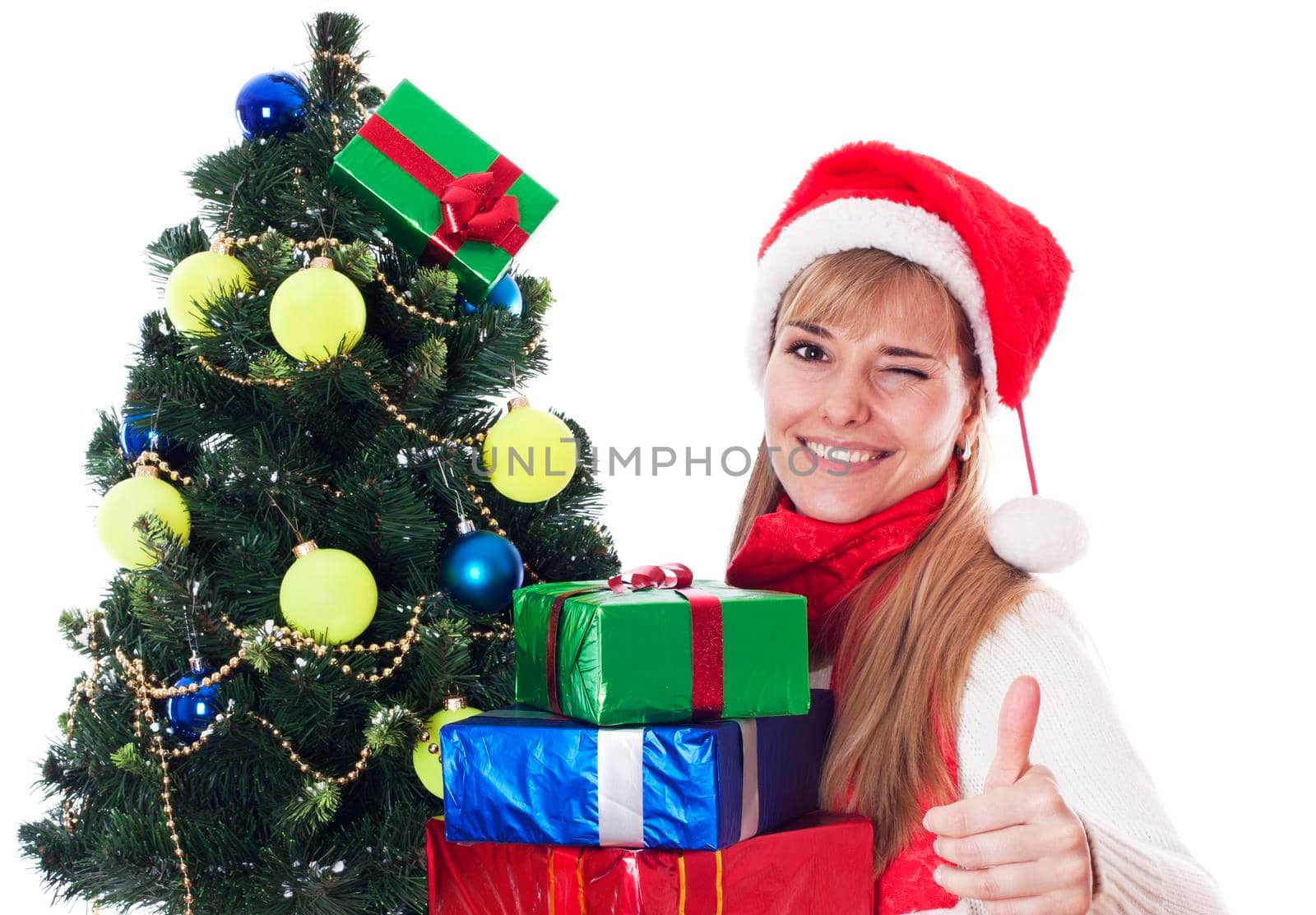 Beautiful young woman with gifts next to christmas tree showing OK sign and winking, isolated on white background.