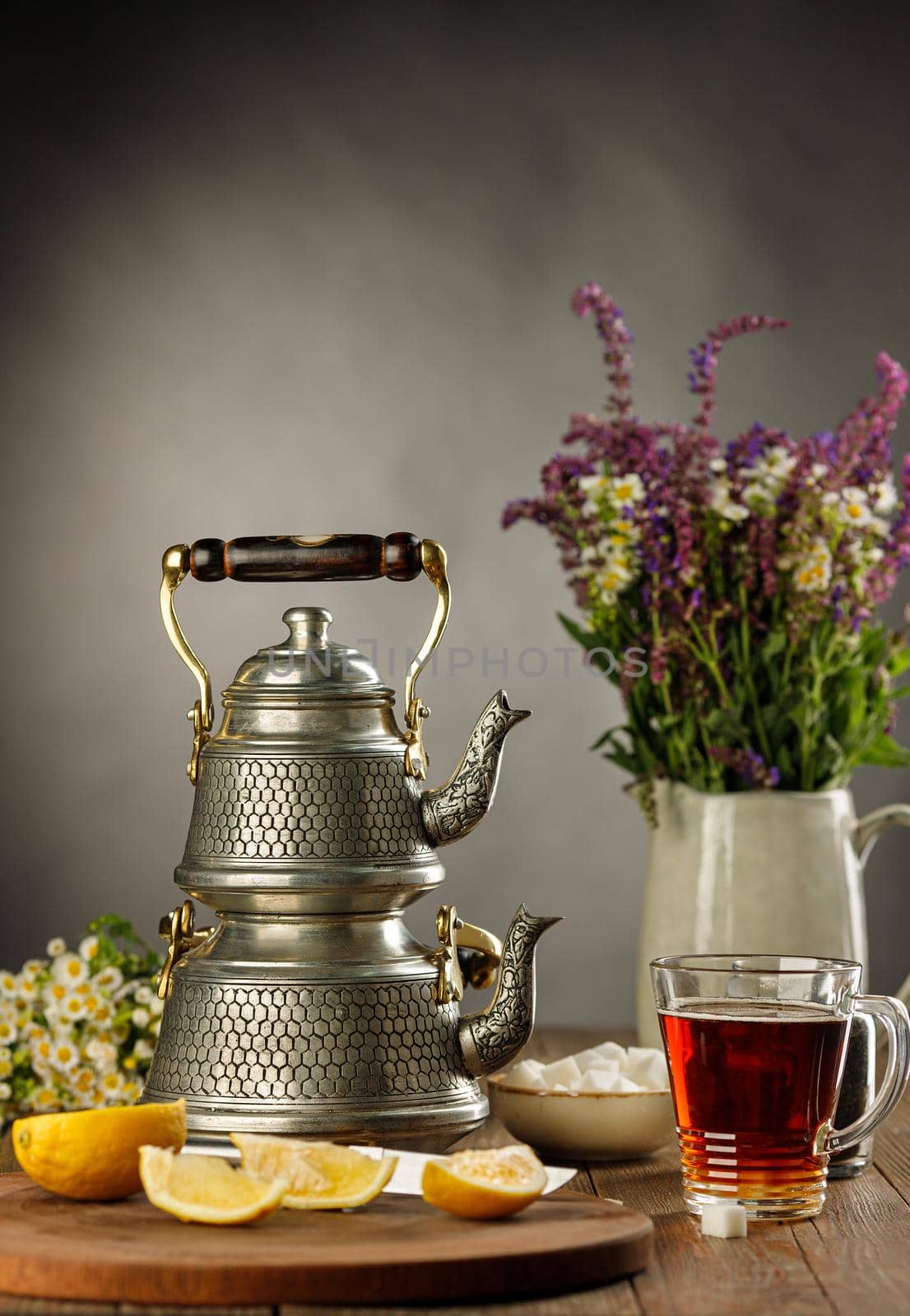Still life tea drinking a cup of tea with a double turkish kettle and a lemon on a wooden board, flowers in the jug by Gravika