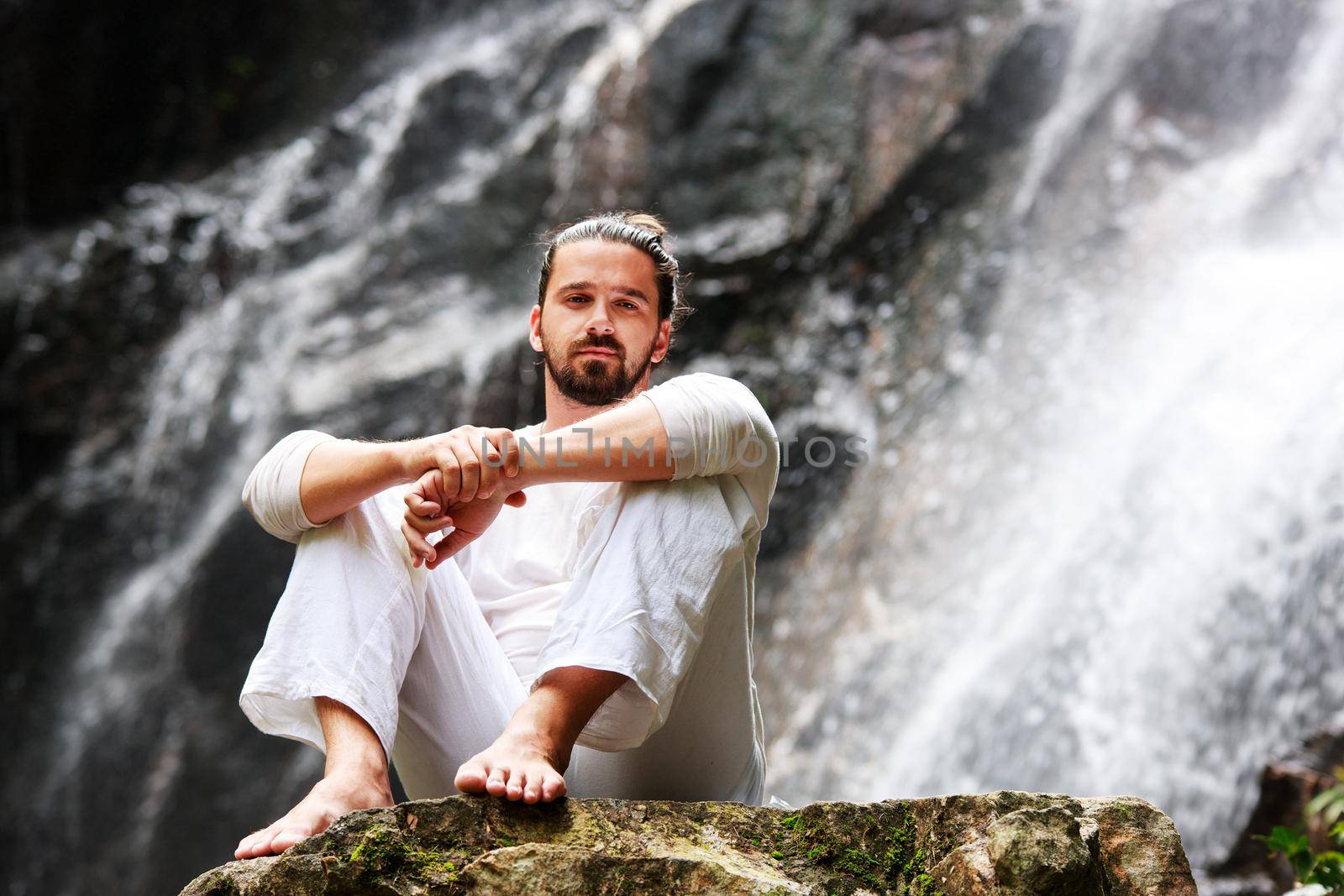 Man sitting in meditation yoga on rock at waterfall in tropical rainforest