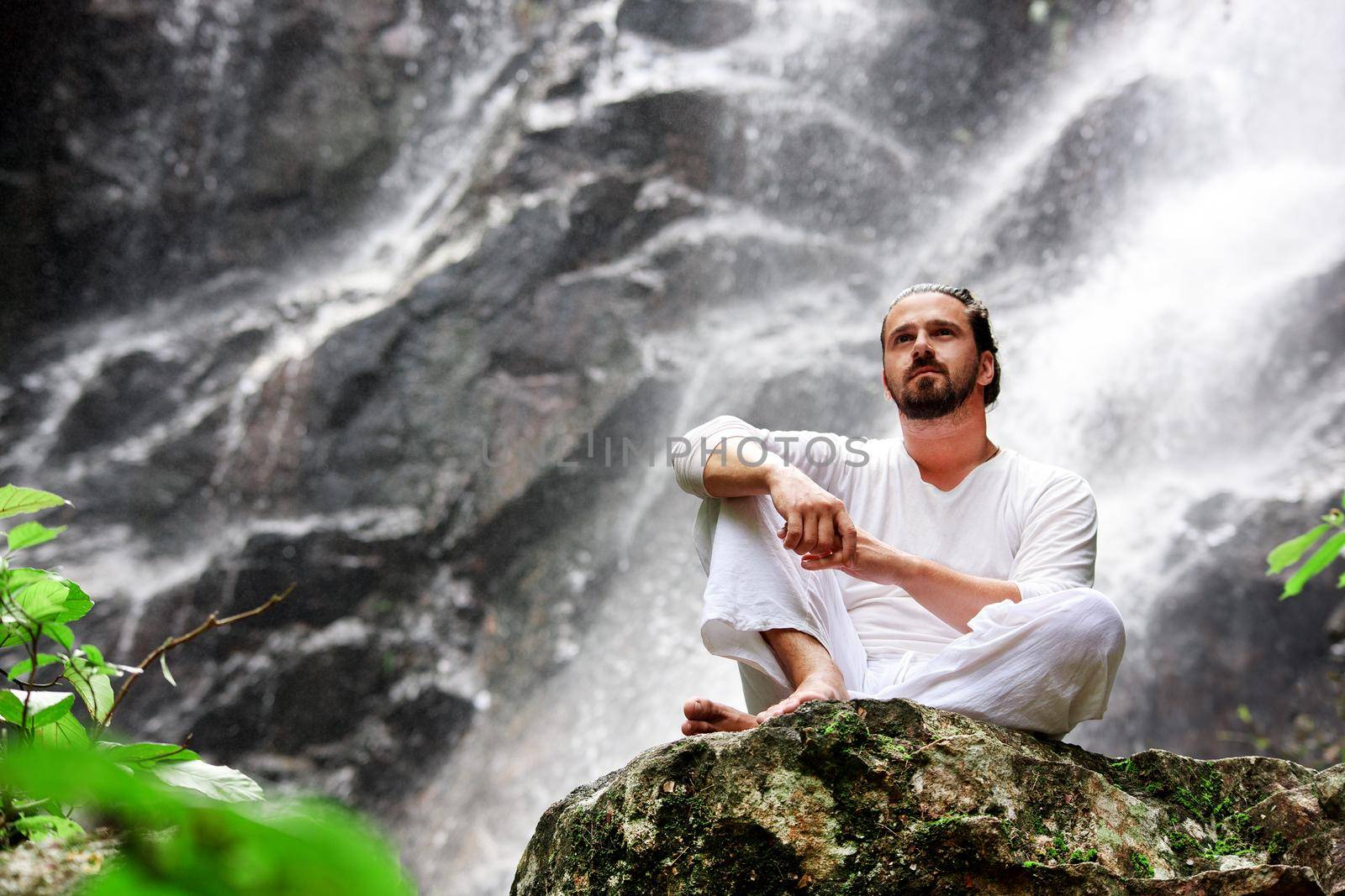 Man sitting in meditation yoga on rock at waterfall in tropical rainforest