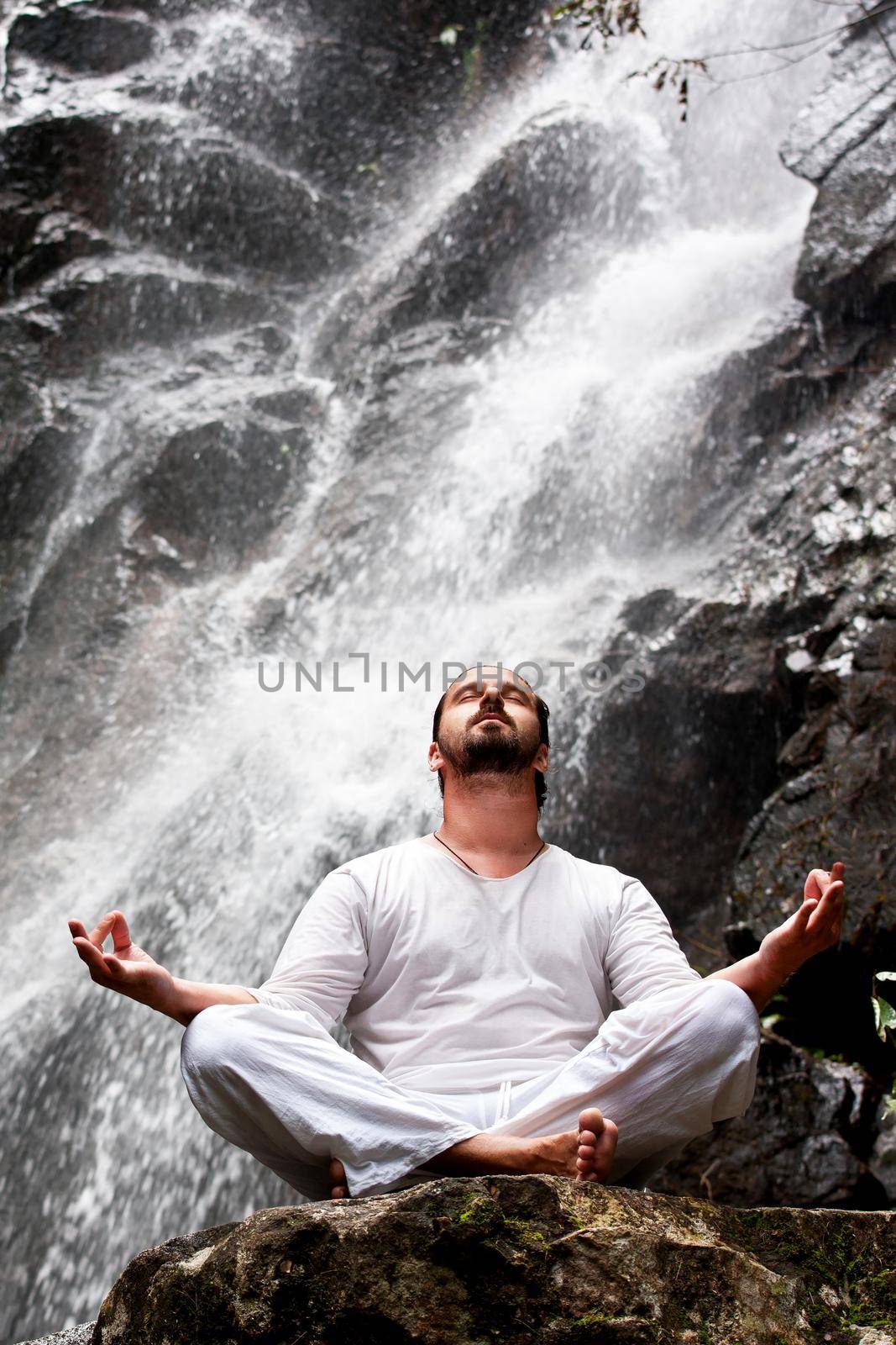 Man sitting in meditation yoga on rock at waterfall in tropical rainforest