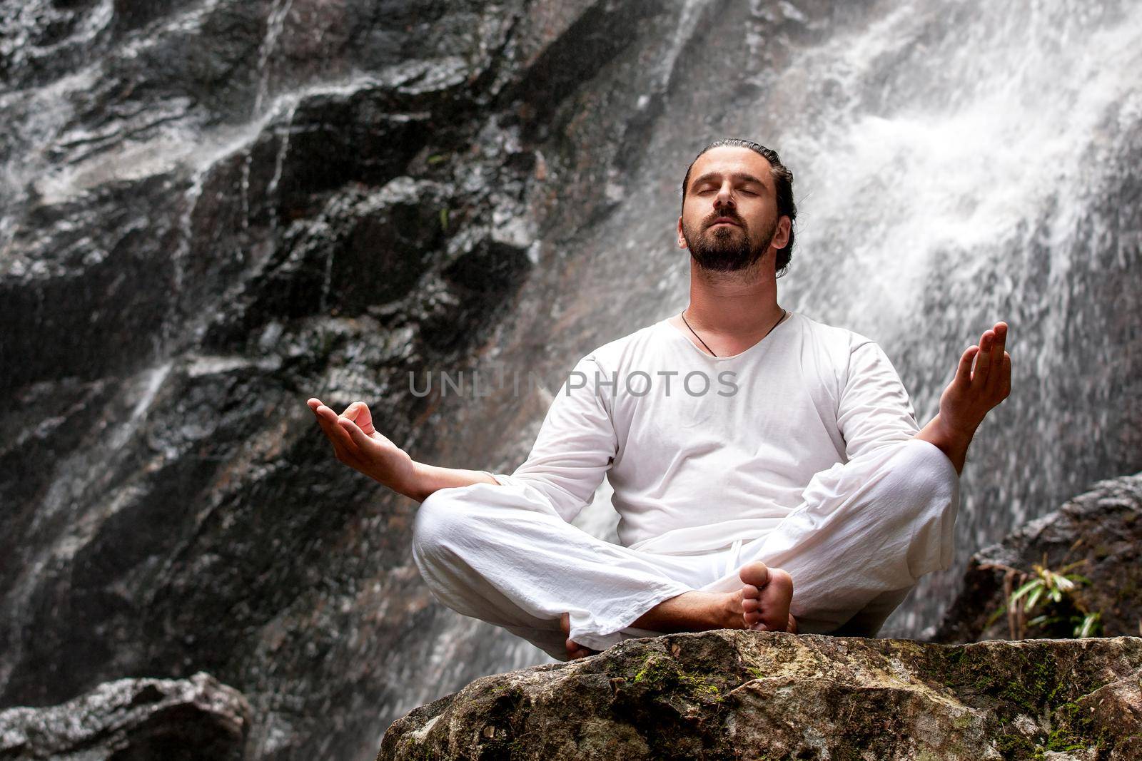 Man sitting in meditation yoga on rock at waterfall in tropical by Jyliana