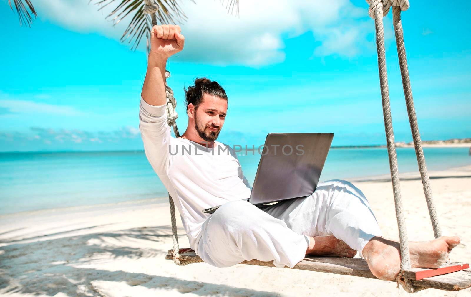 Man working with a laptop, on a hammock in the beach. Concept of digital nomad, remote worker, independent location entrepreneur. Show win