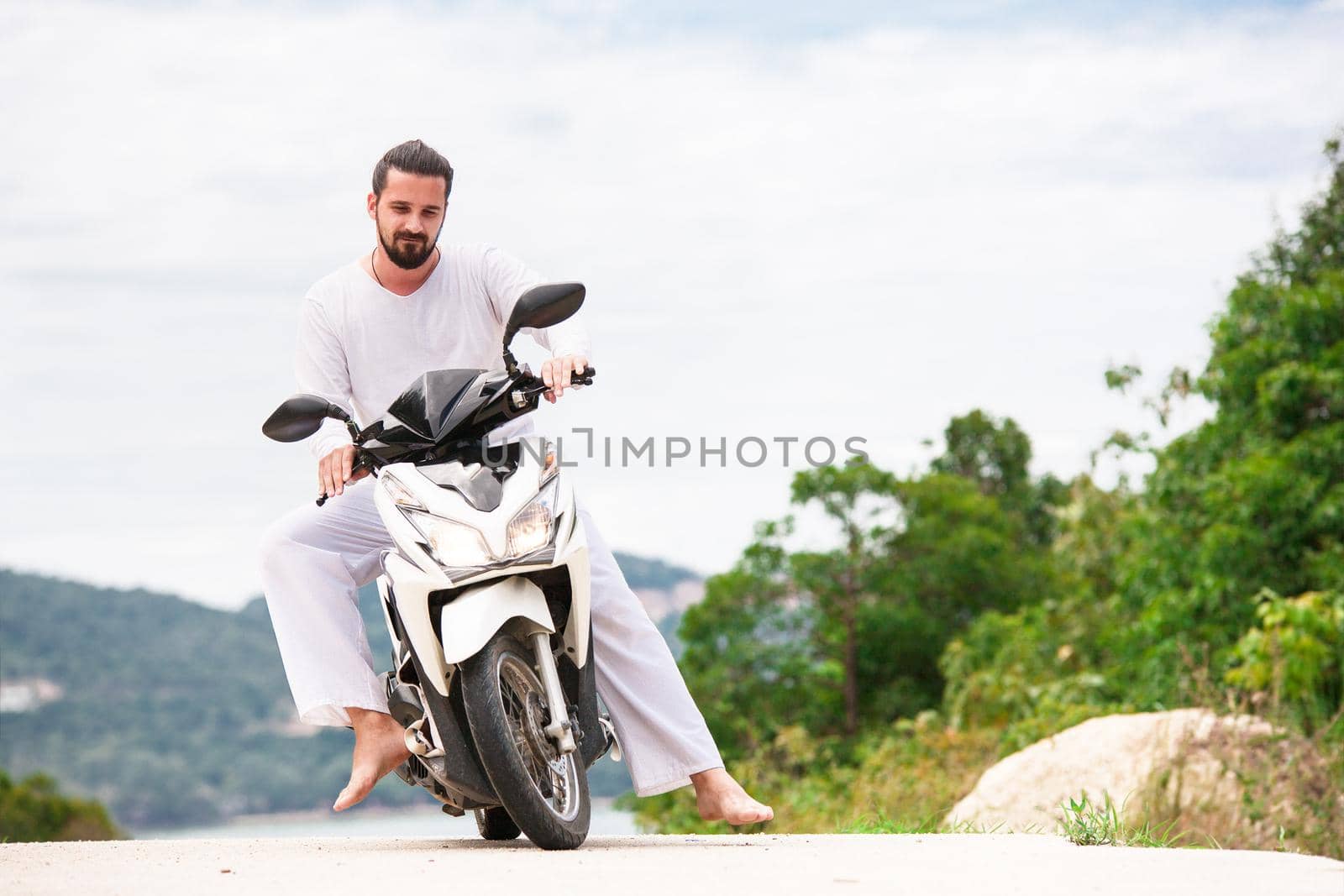 Brutal biker with beard in white sitting on motorbike. Sunny day in the mountains.
