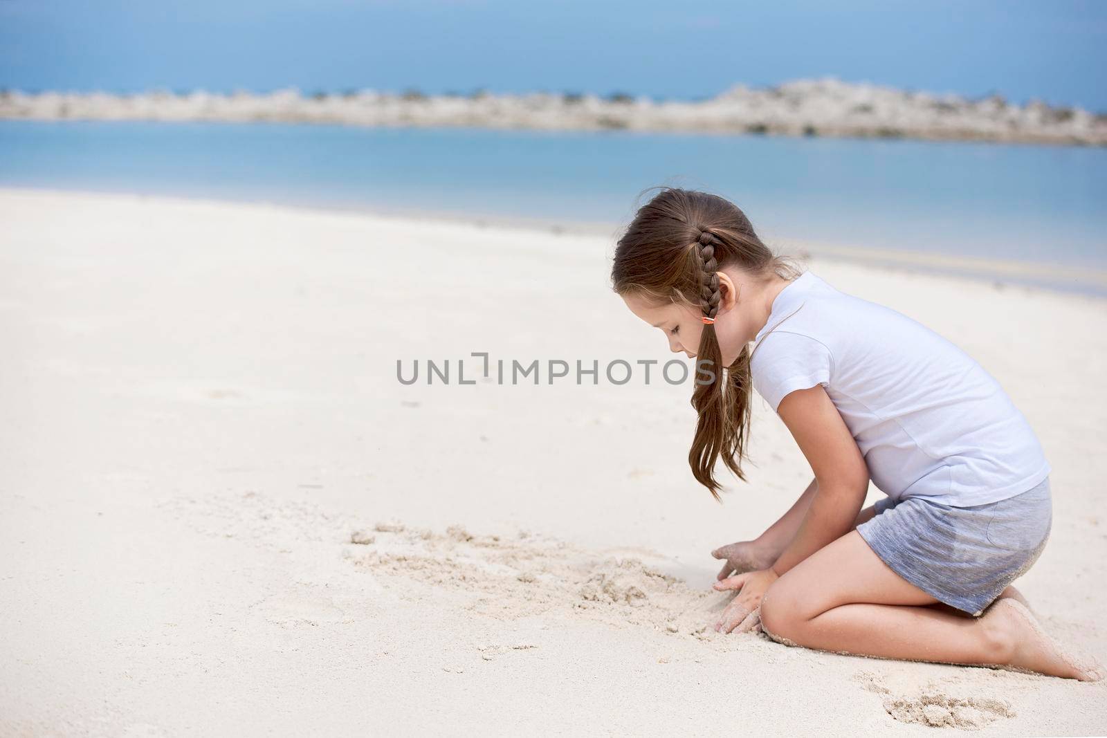 Happy child on the beach. Paradise holiday concept, girl seating on sandy beach with blue shallow water and clean sky by Jyliana