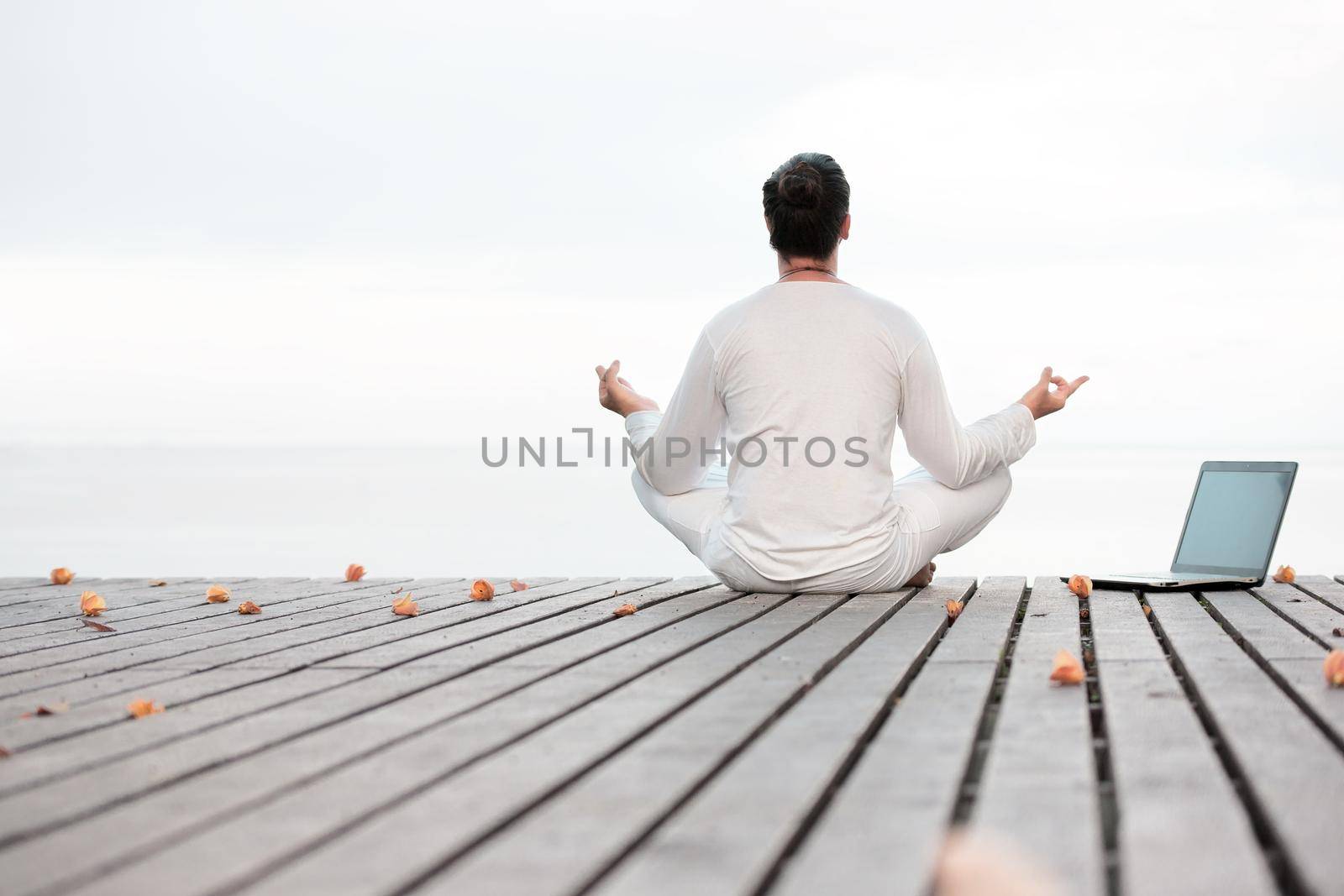 Man in white clothes meditating yoga with laptop on wooden pier by Jyliana